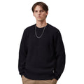 A man wearing a dark navy speckled knit crewneck sweater, paired with khaki pants and a silver chain necklace, photographed against a solid white background.