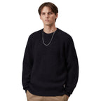 A man wearing a dark navy speckled knit crewneck sweater, paired with khaki pants and a silver chain necklace, photographed against a solid white background.