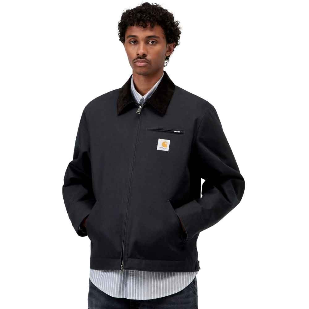 A man wearing a navy Carhartt jacket with a dark brown corduroy collar, a zippered chest pocket, and a visible Carhartt logo patch on the left chest.