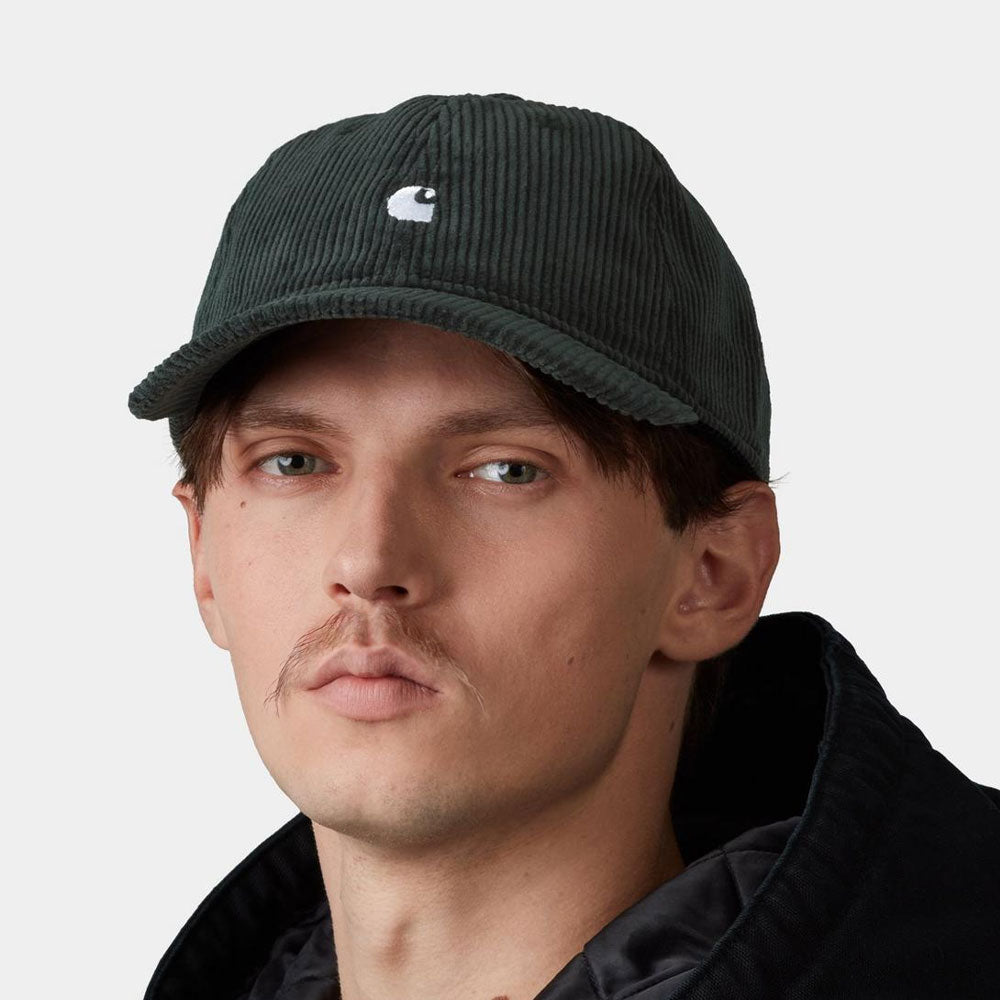A close-up of a man wearing a dark green corduroy baseball cap with a small white Carhartt logo on the front, paired with a black jacket against a light gray background.