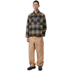 A male model stands against a plain white background wearing a long-sleeve plaid button-up shirt in black, olive green, and tan, paired with tan cargo pants and black shoes.