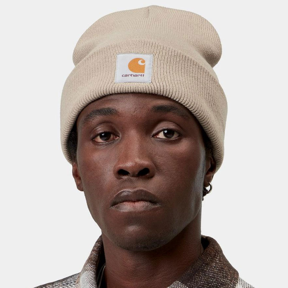 A close-up of a person wearing a beige Carhartt knit beanie with the brand's signature orange and white logo patch on the front, set against a plain light gray background.