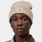 A close-up of a person wearing a beige Carhartt knit beanie with the brand's signature orange and white logo patch on the front, set against a plain light gray background.