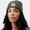 A close-up of a woman wearing a dark olive green Carhartt knit beanie with the signature orange and white logo patch on the front, set against a plain light gray background.
