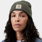 A close-up of a woman wearing a dark olive green Carhartt knit beanie with the signature orange and white logo patch on the front, set against a plain light gray background.