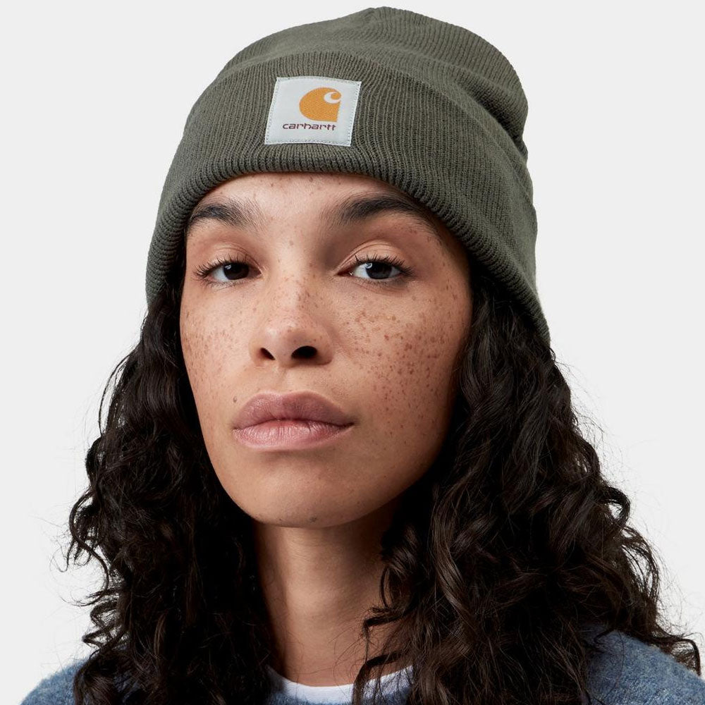 A close-up of a woman wearing a dark olive green Carhartt knit beanie with the signature orange and white logo patch on the front, set against a plain light gray background.