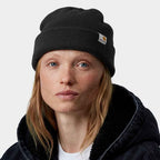 A woman wearing a black Carhartt knit beanie and a dark fleece-lined jacket, photographed against a clean light gray studio background.