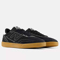 A pair of New Balance low-top sneakers in black suede and canvas with white contrast stitching and a gum rubber sole, shown against a light gray background.