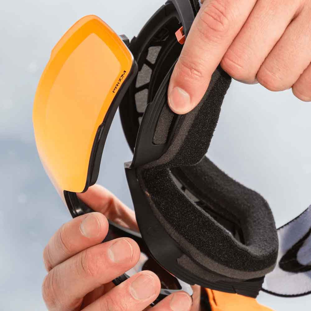 Close-up of a person holding a pair of black ski goggles with an orange-tinted lens and black foam ear pads.