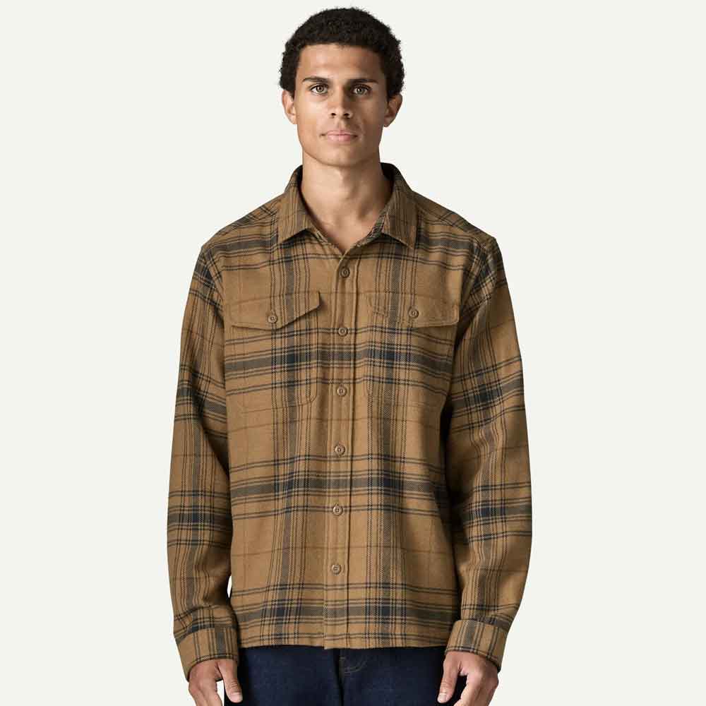 A man wearing a brown and tan plaid button-up shirt with dark navy lines, paired with dark blue jeans, photographed against a solid off-white background.