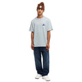 A man modeling a light blue t-shirt with a small purple and white mountain logo on the left chest, paired with dark blue jeans, white sneakers, and a brown knit beanie hat, set against a plain white background.