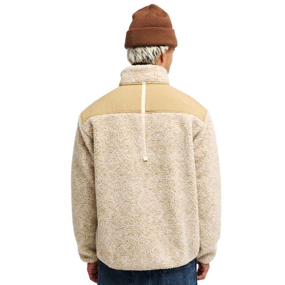 A man viewed from behind wearing a fleece pullover jacket with a tan yoke and a cream sherpa body, paired with a brown beanie and blue jeans against a white background.