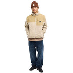 A model wearing a beige and cream fleece zip-up jacket, a brown beanie, a tan shirt, blue jeans, and black shoes, standing against a solid white background.