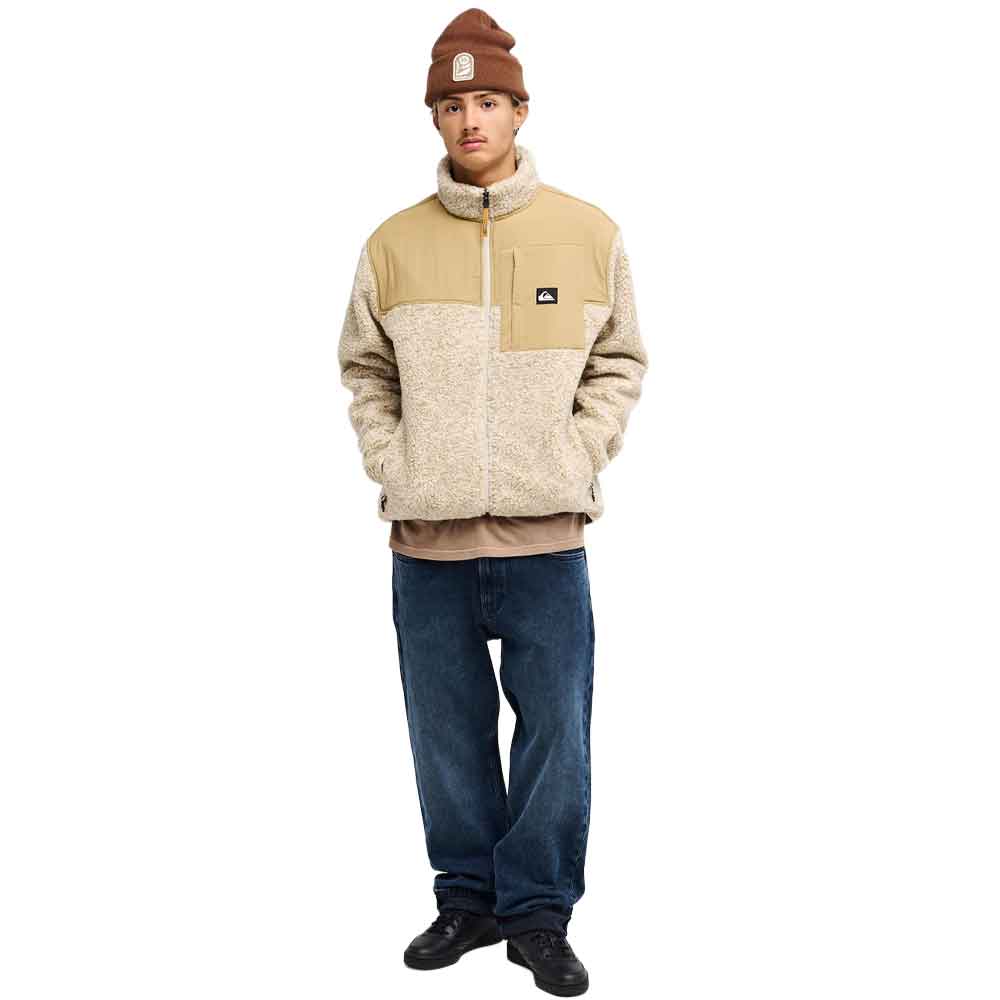A model wearing a beige and cream fleece zip-up jacket, a brown beanie, a tan shirt, blue jeans, and black shoes, standing against a solid white background.