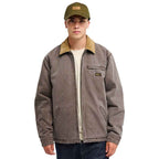 A man wearing a brown work jacket with a tan corduroy collar, an olive green baseball cap, and a cream t-shirt, photographed against a solid white background.