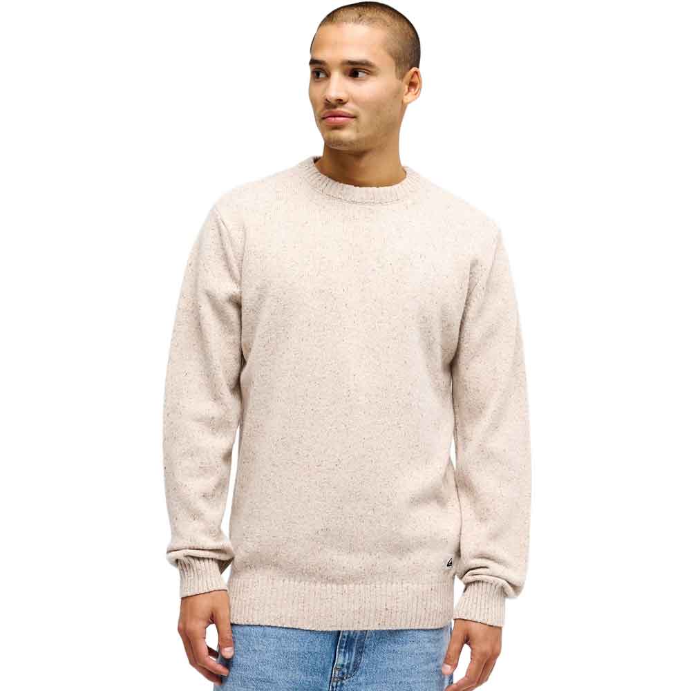 A man wearing a light beige knit crewneck sweater and blue jeans, standing against a plain white background.