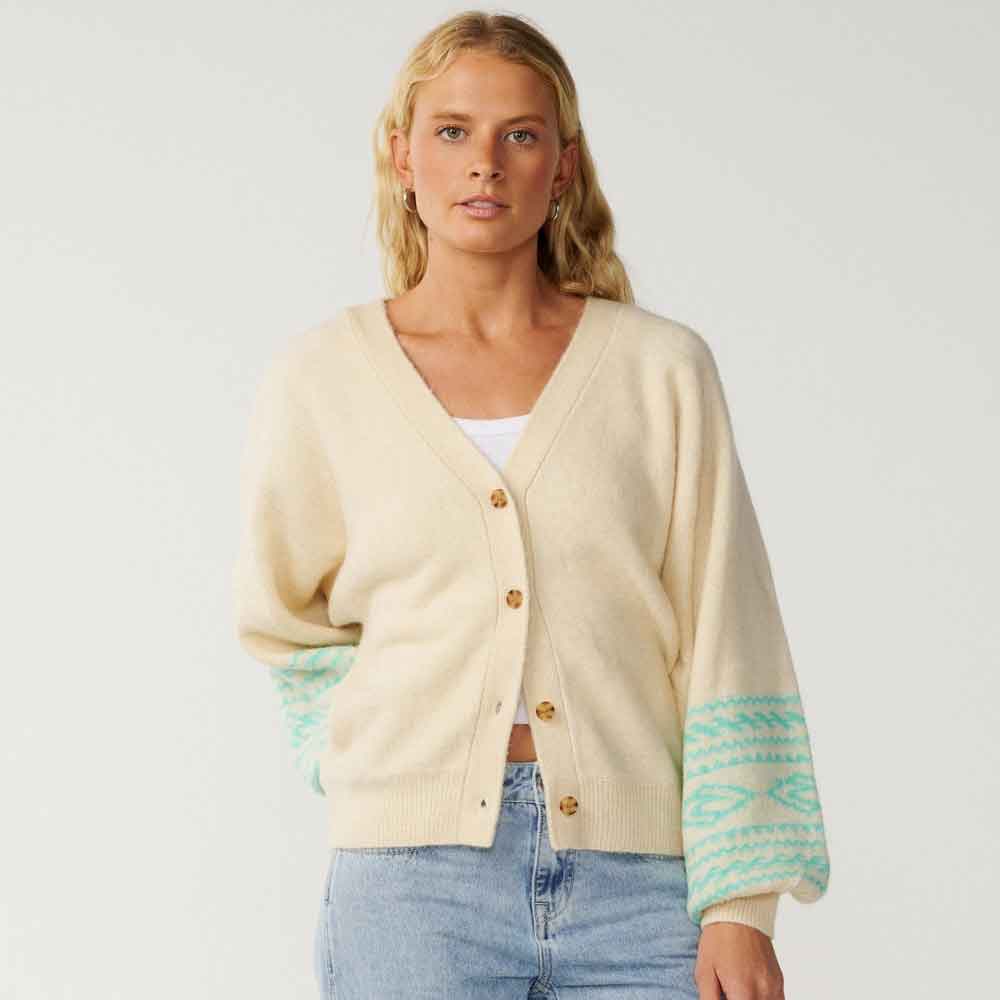 A woman modeling a cream-colored cardigan with mint green patterned cuffs and wooden buttons, paired with light blue jeans against a plain light gray background.