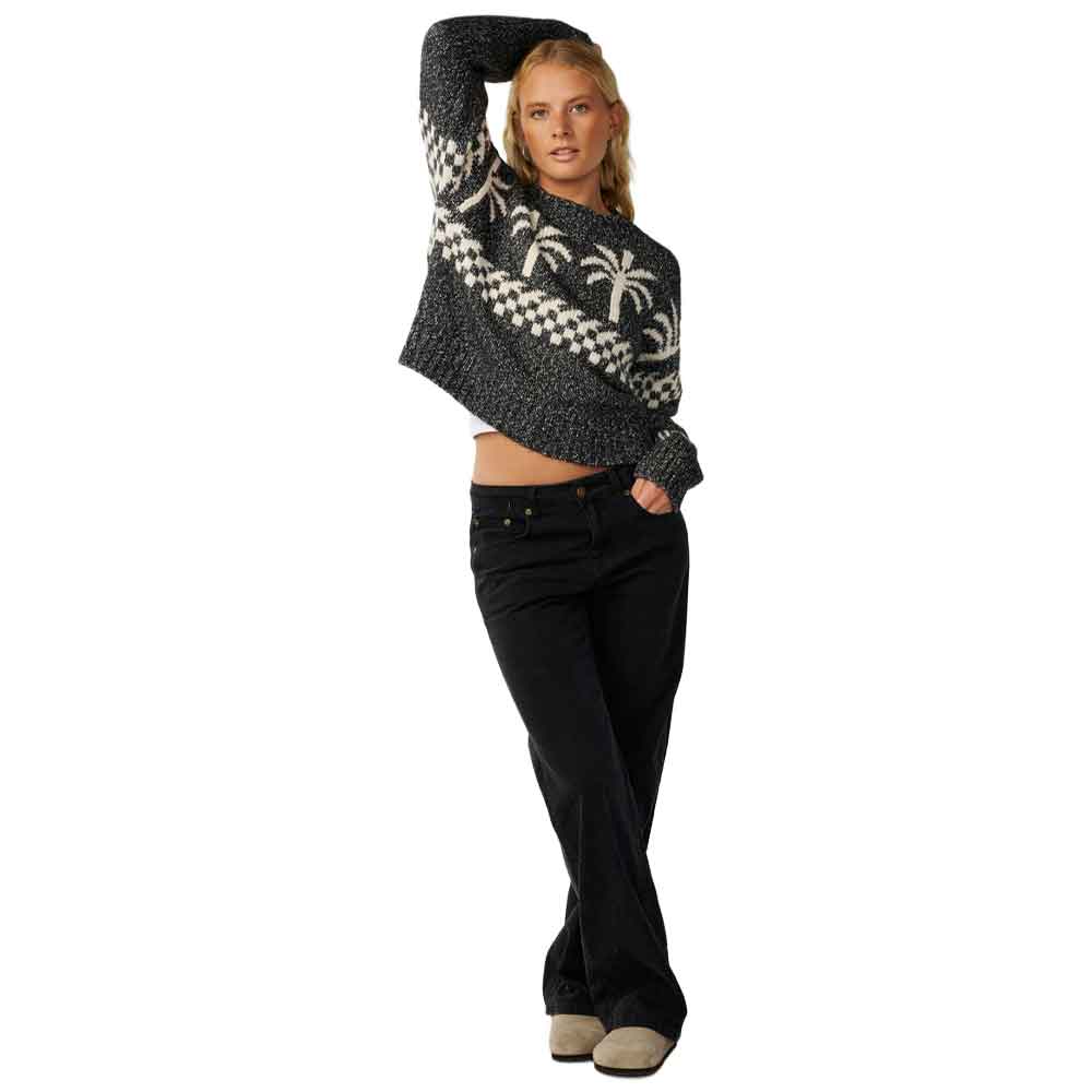 A woman modeling a cropped knit sweater with a black and white geometric pattern and white palm tree motifs, paired with black jeans and beige slip-on shoes, against a plain white background.