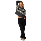 A woman modeling a cropped knit sweater with a black and white geometric pattern and white palm tree motifs, paired with black jeans and beige slip-on shoes, against a plain white background.