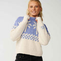 A woman wearing a cream and blue knit sweater with a Fair Isle pattern and ribbed hem, paired with black jeans, against a light gray studio background.