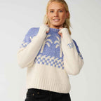 A woman wearing a cream and blue knit sweater with a Fair Isle pattern and ribbed hem, paired with black jeans, against a light gray studio background.