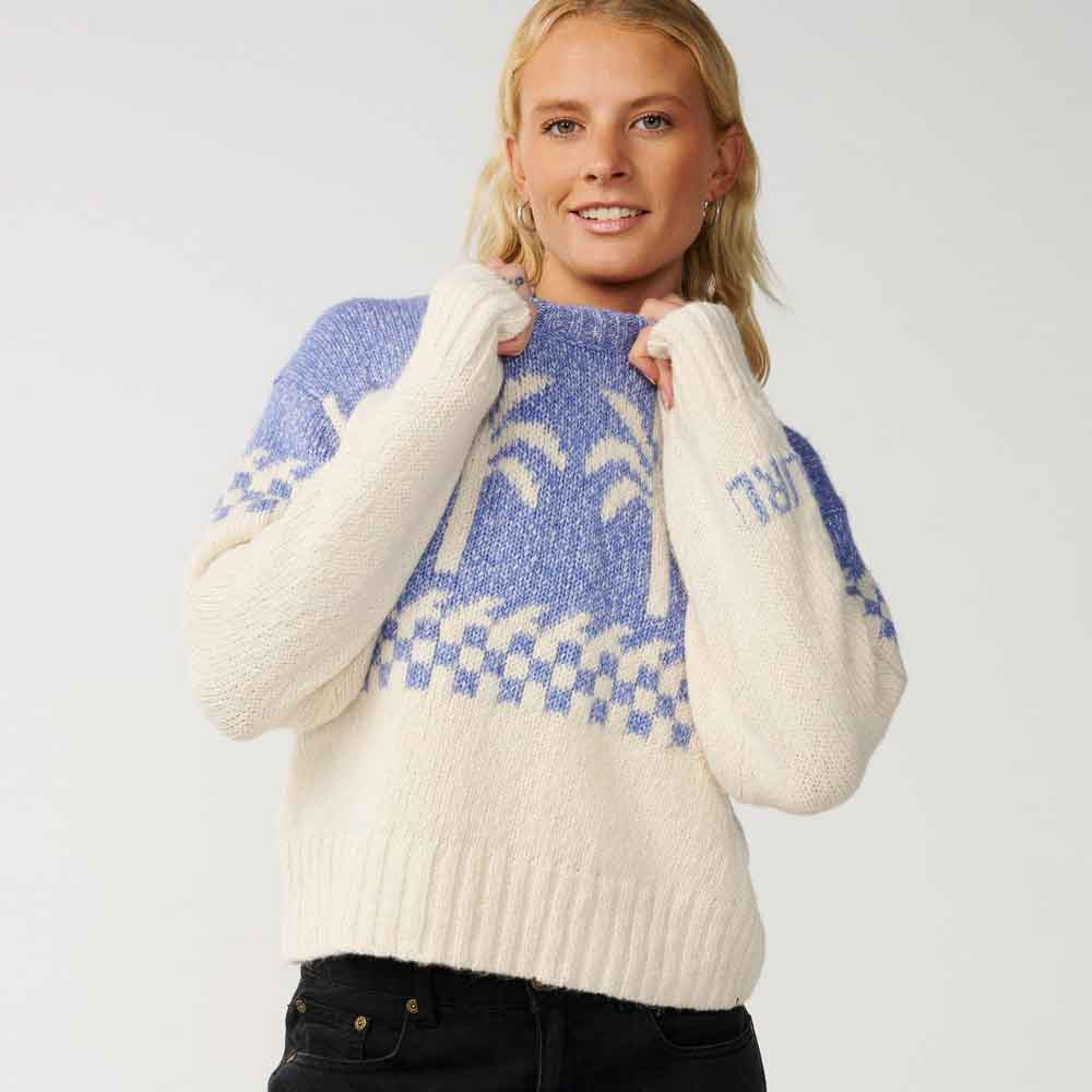 A woman wearing a cream and blue knit sweater with a Fair Isle pattern and ribbed hem, paired with black jeans, against a light gray studio background.