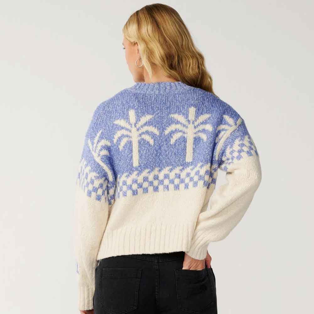 A woman shown from the back wearing a cream and light blue knit sweater with a palm tree pattern, paired with black jeans, against a plain light gray studio background.