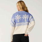 A woman shown from the back wearing a cream and light blue knit sweater with a palm tree pattern, paired with black jeans, against a plain light gray studio background.