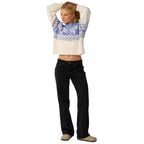 A woman modeling a cream knit sweater with a blue and white Nordic pattern across the chest and sleeves, paired with black jeans and beige slip-on shoes, against a plain white background.