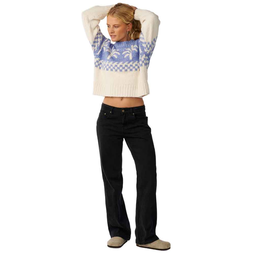 A woman modeling a cream knit sweater with a blue and white Nordic pattern across the chest and sleeves, paired with black jeans and beige slip-on shoes, against a plain white background.