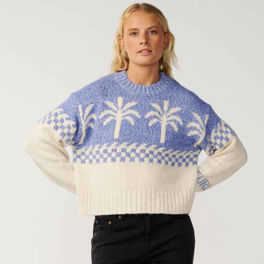 A blonde woman wearing a blue and cream knit sweater with palm tree patterns, paired with black jeans, against a light gray studio background.