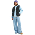 A model wearing a light blue hoodie, a black textured sleeveless vest, wide-leg light blue jeans, a purple beanie, and tan slip-on shoes, standing against a plain white background.