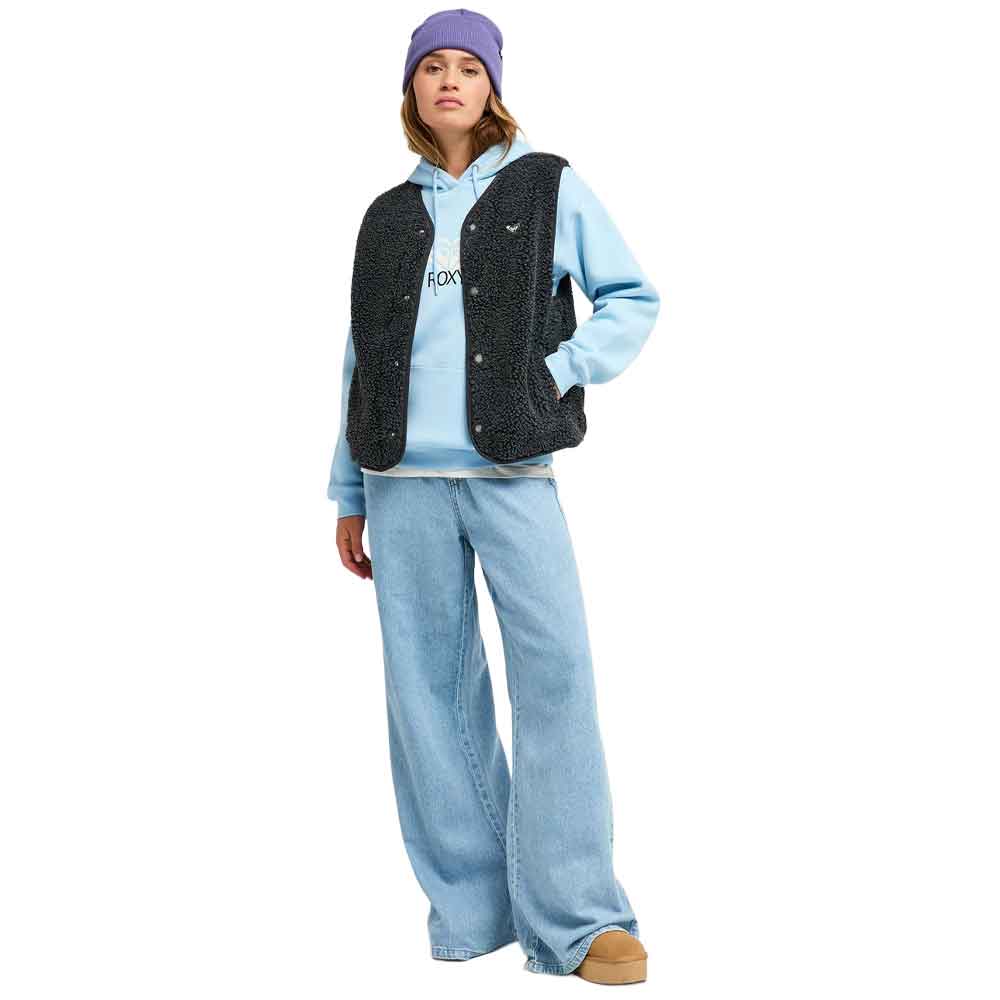 A model wearing a light blue hoodie, a black textured sleeveless vest, wide-leg light blue jeans, a purple beanie, and tan slip-on shoes, standing against a plain white background.