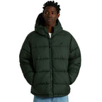 A man wearing a dark forest green hooded puffer jacket, styled with a white t-shirt and blue jeans, photographed against a solid white background.