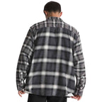 A man viewed from behind wearing a dark gray and white plaid flannel shirt jacket, paired with black pants, against a solid white background.