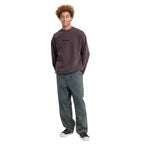 A model wearing a dark brown crewneck sweatshirt with a small black 'uniqlo' logo on the chest, paired with dark gray relaxed-fit jeans and black sneakers, standing against a solid white background.