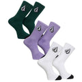 Three pairs of Volcom crew socks in forest green, light purple, and white, arranged diagonally on a solid white background. Each pair features the signature Volcom mountain logo embroidered on the upper cuff.