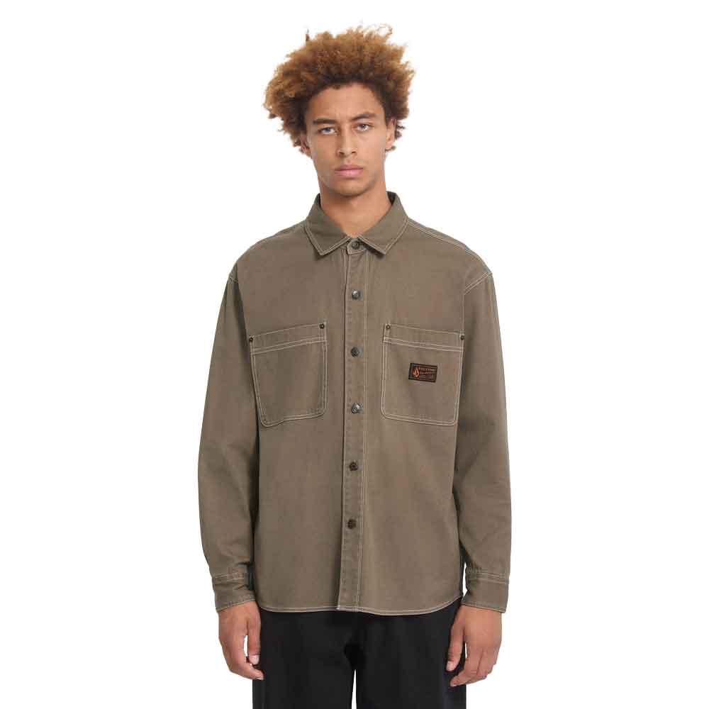 A model with curly light brown hair wearing a muted brown utility shirt jacket with two chest pockets and a small brand patch on the left chest, paired with black pants against a solid white background.