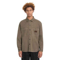 A model with curly light brown hair wearing a muted brown utility shirt jacket with two chest pockets and a small brand patch on the left chest, paired with black pants against a solid white background.