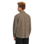 A man with curly brown hair is shown from the back wearing an oversized, light brown utility shirt and black pants against a solid white background.
