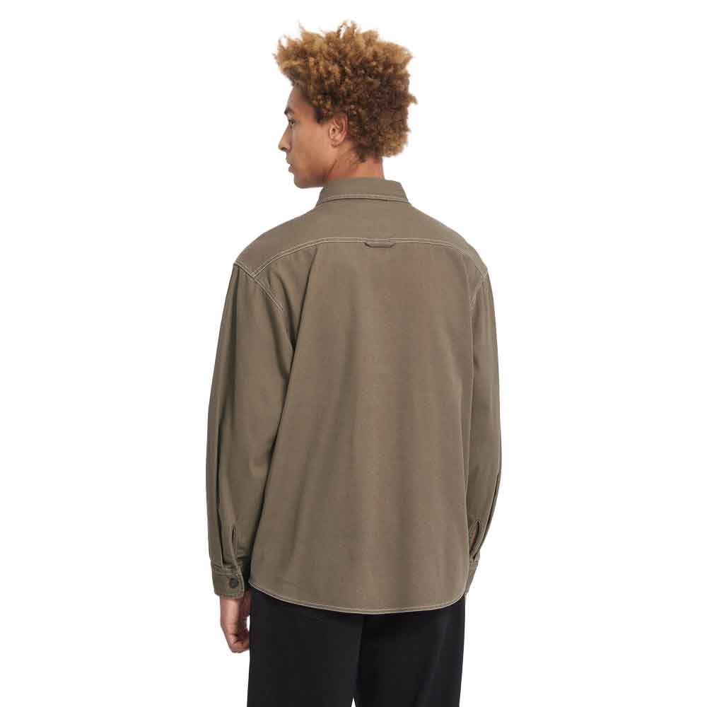 A man with curly brown hair is shown from the back wearing an oversized, light brown utility shirt and black pants against a solid white background.