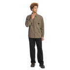 A man modeling a muted olive-brown long-sleeve button-up shirt with a chest pocket, paired with black pants and dark shoes, against a plain white background.