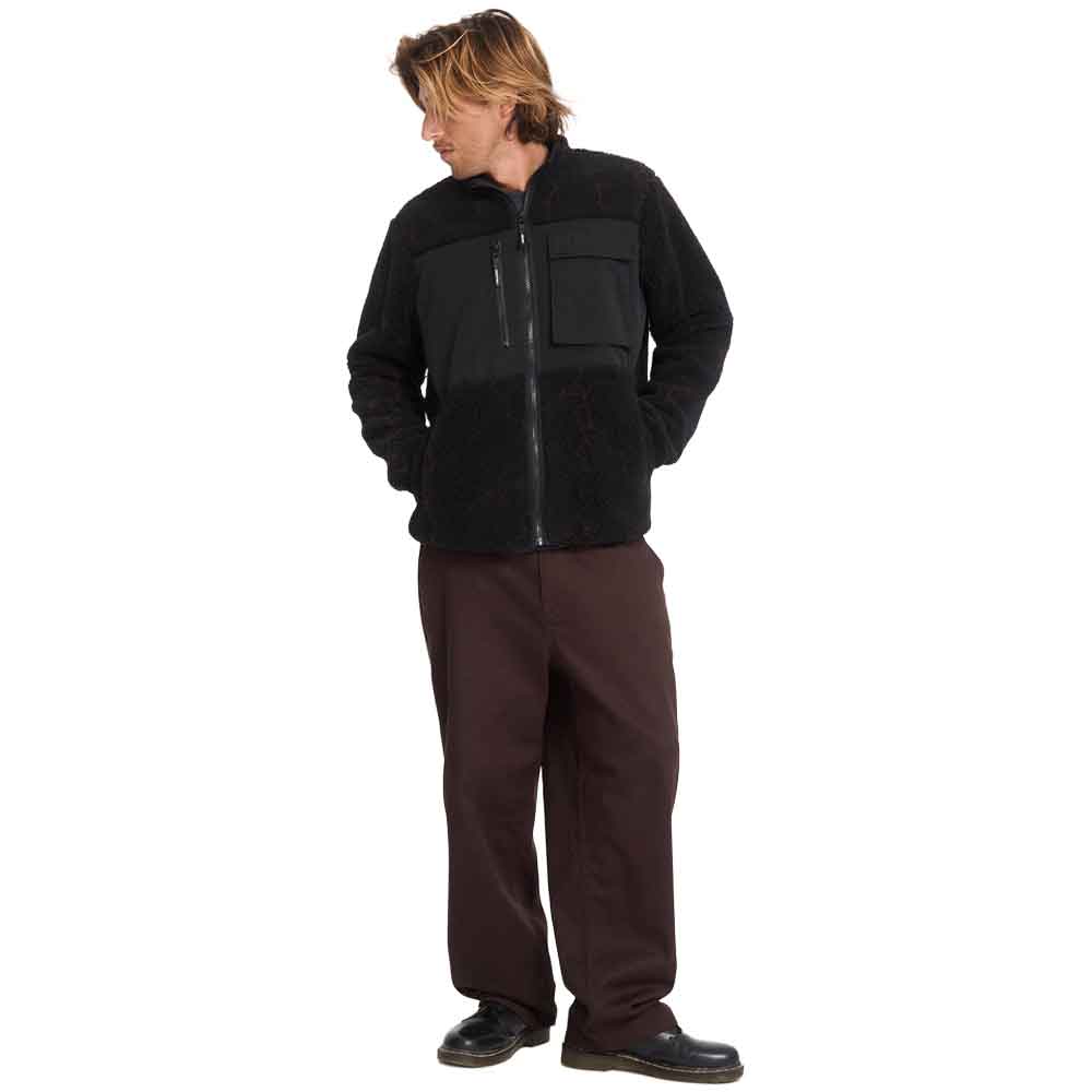 A man wearing a black fleece zip-up jacket with a chest pocket and dark brown pants, standing against a plain white background.