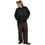 A man wearing a black fleece zip-up jacket with a chest pocket and dark brown pants, standing against a plain white background.