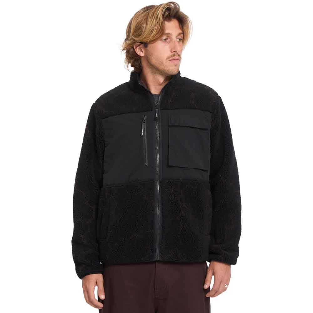 A man wearing a black fleece jacket with a zippered chest pocket and ribbed cuffs, paired with dark brown pants, photographed against a solid white background.