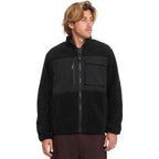 A man wearing a black fleece jacket with a zippered chest pocket and ribbed cuffs, paired with dark brown pants, photographed against a solid white background.
